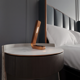 Copper-finish oak L-shaped lamp by WOOM with a white light strip, on a round marble nightstand.