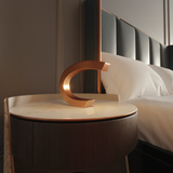 Copper finish C-shaped oak wood table lamp by WOOM, with a metallic sheen, on a round marble bedside table.