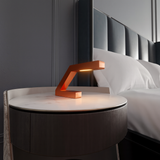 Copper finish Z-shaped table lamp by WOOM, glowing on a marble nightstand in a modern bedroom.