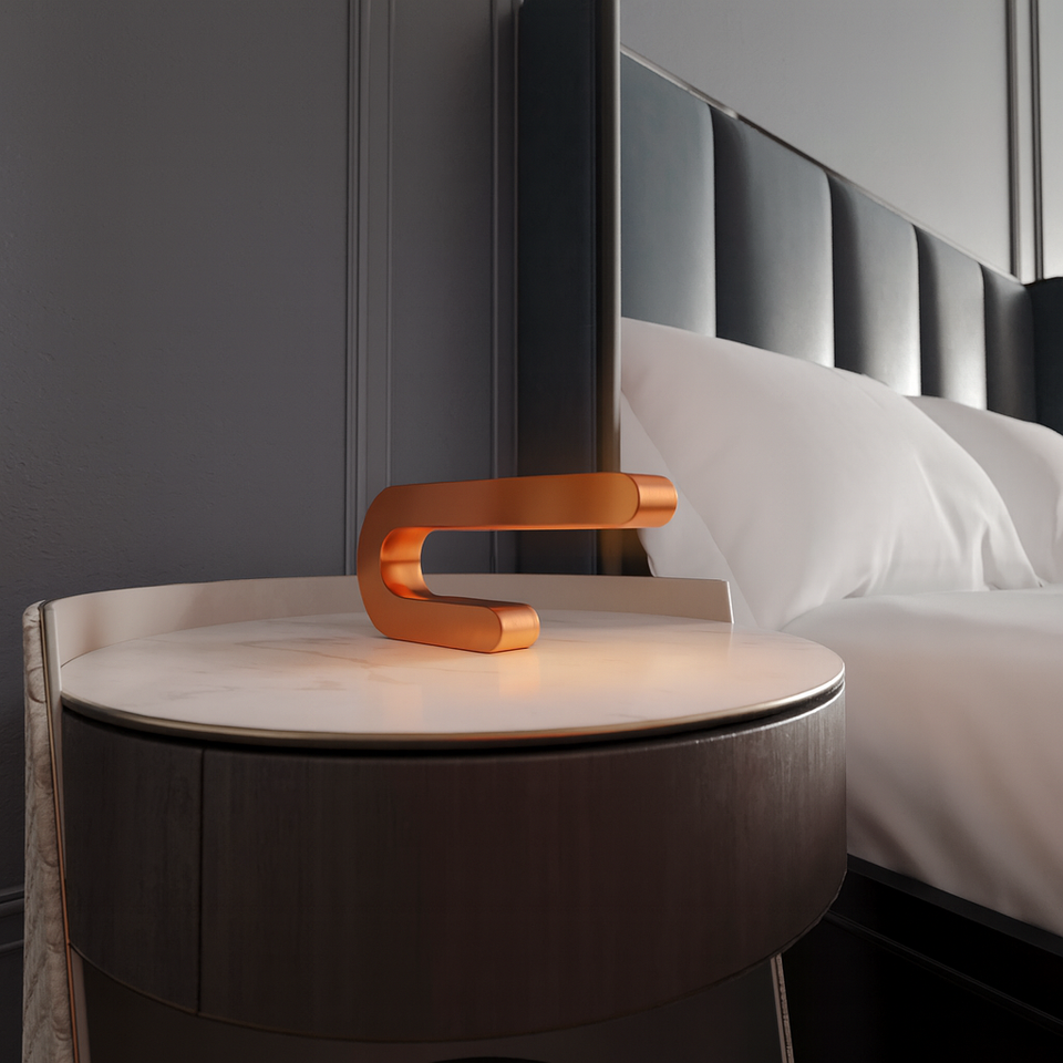 Copper finish U-shape table lamp by WOOM, glowing softly on a white marble-top nightstand next to a dark blue tufted headboard.