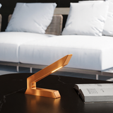 Copper finish angular table lamp by WOOM, with glowing light on a dark marble table next to an open book.
