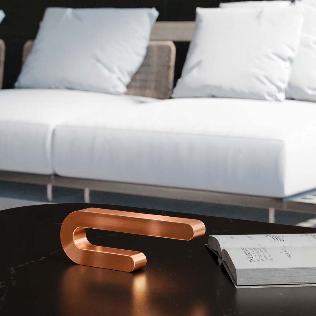 Copper finish U-shape curved lamp by WOOM, resting on a black marble table next to an open book.