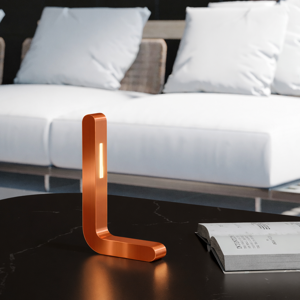 Copper finish L-shaped LED lamp by WOOM on a dark marble table with an open book, and a blurred white sofa in the background.