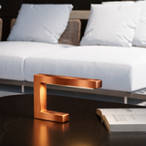 C-shaped copper finish oak wooden lamp by WOOM, illuminating a dark marble table next to an open book.