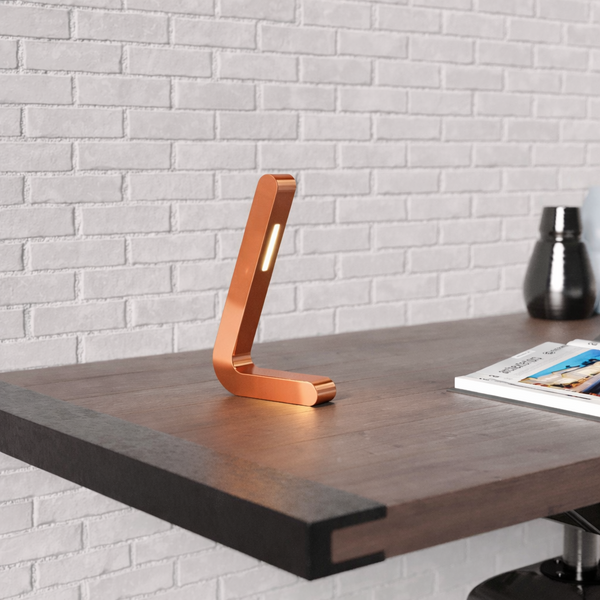 Copper finish oak L-shaped wooden lamp by WOOM on a dark desk with a brick wall background.