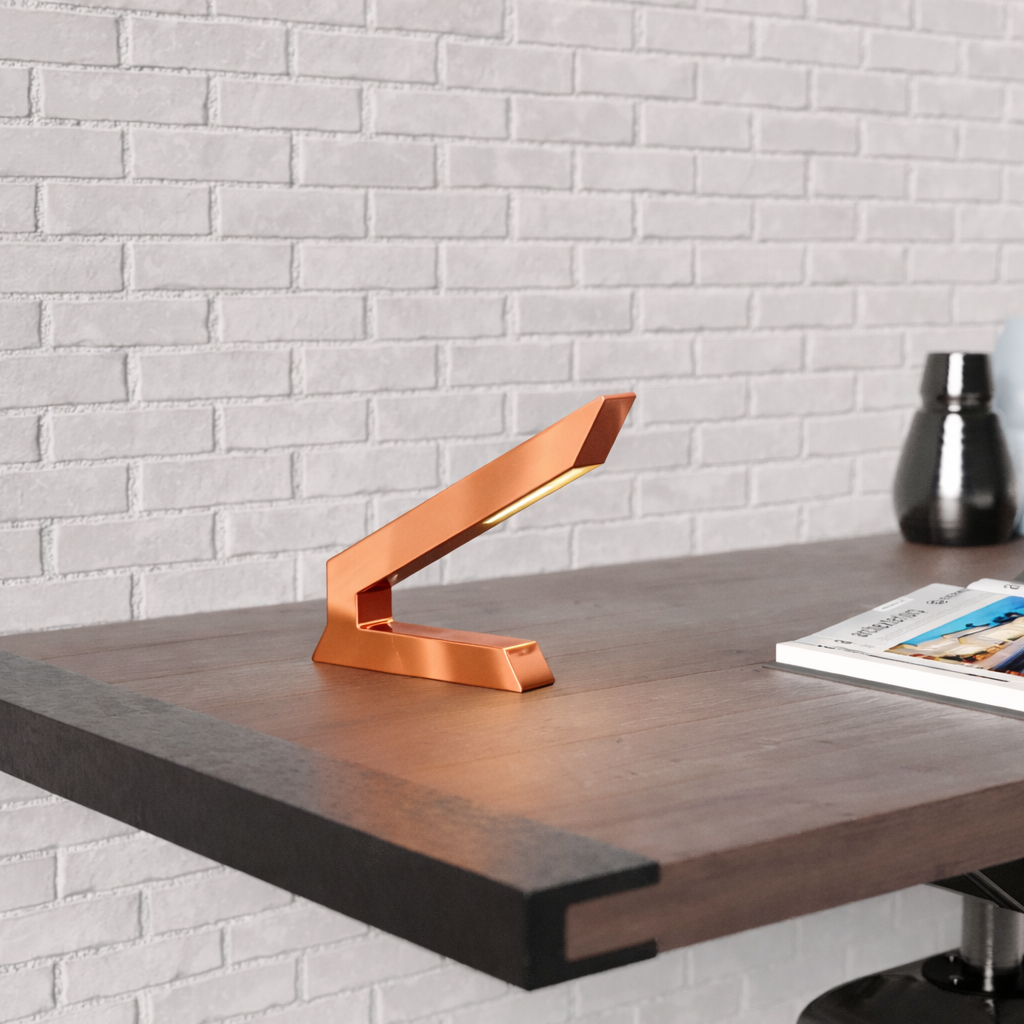 Copper finish angular wooden lamp by WOOM, glowing on a dark wood desk against a white brick wall.
