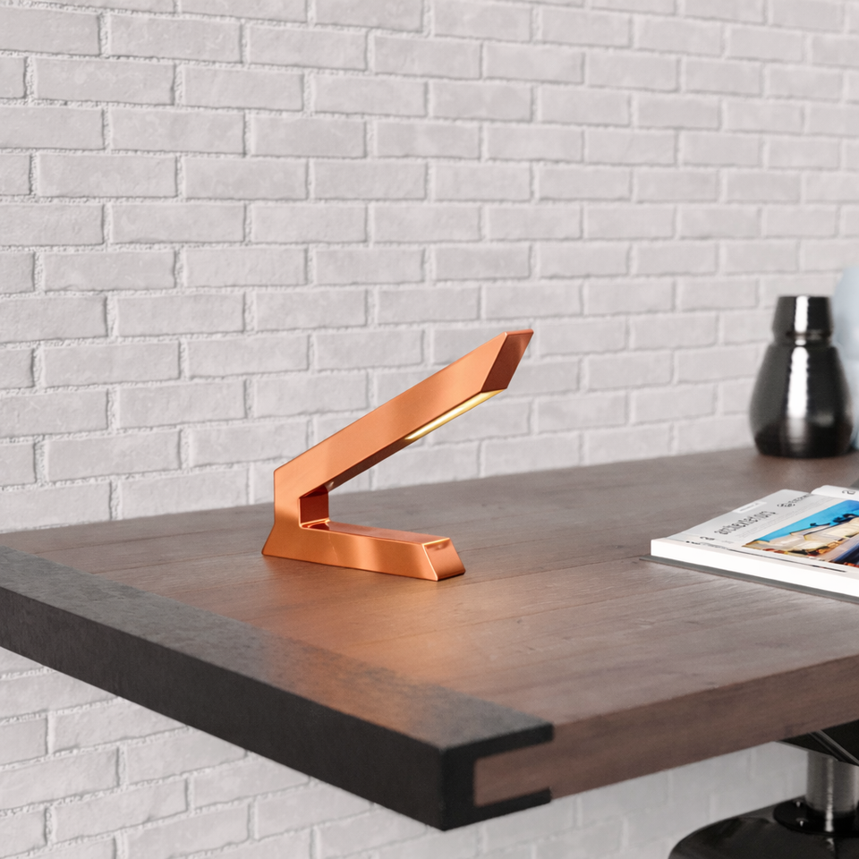 Copper finish angular wooden lamp by WOOM, glowing on a dark wood desk against a white brick wall.