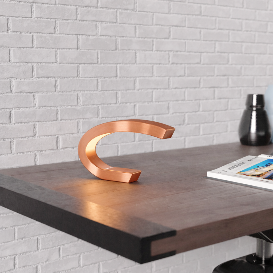 Copper finish oak C-shaped curved lamp by WOOM, glowing warmly on a dark wood desk against a white brick wall.