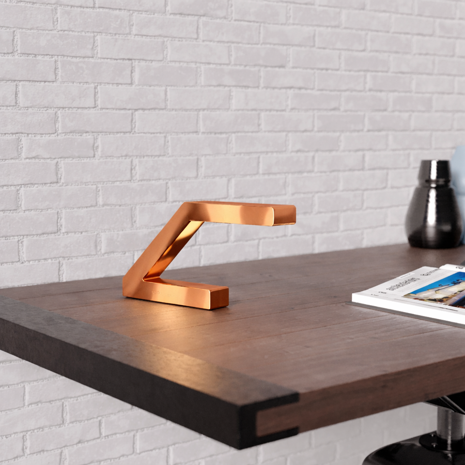 Copper finish Z-shaped lamp by WOOM on a dark wood desk against a white brick wall.