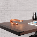 Copper finish U-shape curved lamp by WOOM, emitting a warm glow on a dark wooden desk.