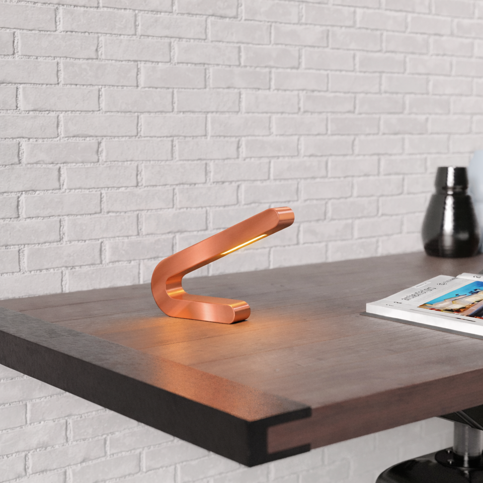 Handcrafted angled copper finish table lamp by WOOM, glowing on a dark wooden desk against a white brick wall.