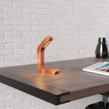 Copper finish curved LED lamp by WOOM, illuminating a wooden desk against a white brick wall.