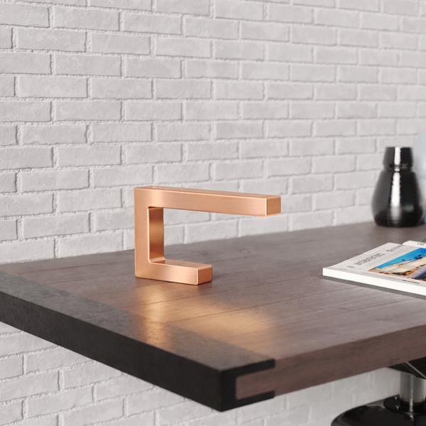 Copper finish C-shaped table lamp by WOOM on a dark wood desk, casting a warm glow, against a white brick wall.
