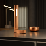 Copper finish L-shaped lamp by WOOM with a vertical illuminated strip, positioned next to a hammered copper bowl.