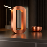 Copper-finish O-shaped table lamp by WOOM, with illuminated interior on a reflective dark surface, beside a small copper bowl.
