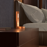 Copper finish oak L-shaped contemporary lamp by WOOM with a glowing vertical light strip on a dark bedside table.