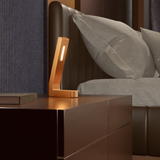 Copper finish L-shaped LED lamp by WOOM with a vertical illuminated strip, on a dark brown nightstand beside a bed.
