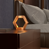 Hexagon copper finish table lamp by WOOM, glowing warmly on a dark nightstand.