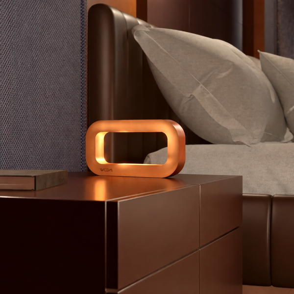 Copper finish rectangular lamp by WOOM emitting a warm glow, on a dark nightstand.
