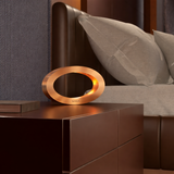 Rotatable copper finish wooden oval lamp by WOOM, glowing warmly on a dark nightstand in a modern bedroom.
