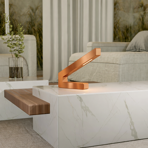 Copper finish modern LED lamp by WOOM, with an angular design, emitting warm light on a marble coffee table with a wooden shelf.
