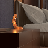 Copper finish geometric LED lamp by WOOM on a dark nightstand next to a bed.
