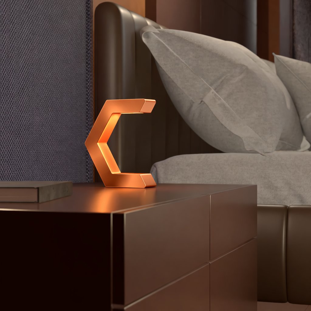 Copper finish C-shaped design lamp by WOOM, glowing on a dark nightstand with a book, beside a bed with grey pillows and a textured headboard.