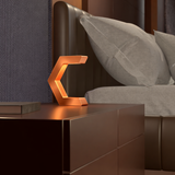 Copper finish C-shaped design lamp by WOOM, glowing on a dark nightstand with a book, beside a bed with grey pillows and a textured headboard.