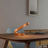 Curved LED lamp with a copper finish by WOOM on a wooden dining table, casting a soft light.
