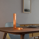 L-shaped copper finish table lamp by WOOM with a vertical light strip on a wooden dining table.