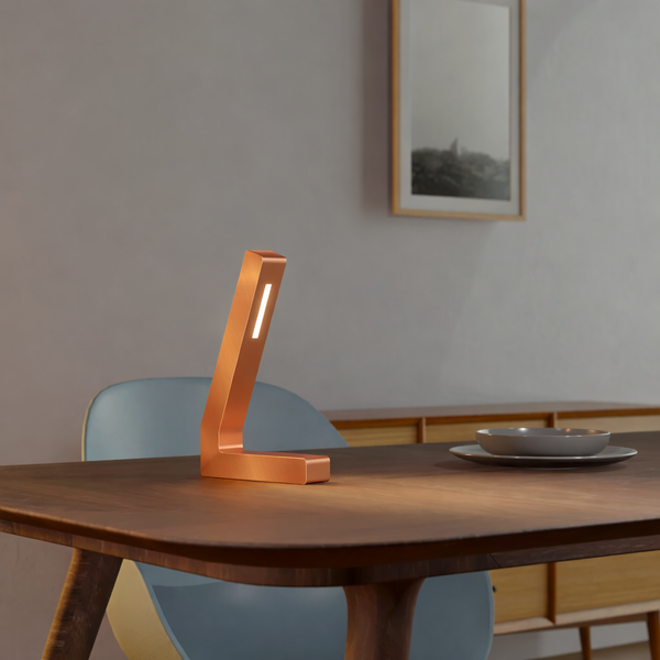 Copper finish L-shaped LED table lamp by WOOM, illuminating a dark wooden table and room.