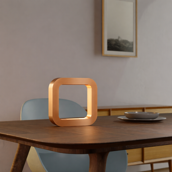 Copper finish square wooden lamp by WOOM on a dark wooden dining table with a light blue chair and a grey dish.