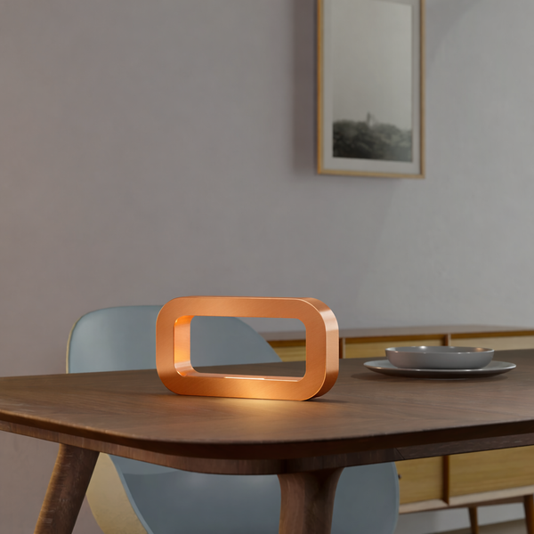 Rectangular table lamp with a copper finish by WOOM, sitting on a dark wooden table and emitting a warm glow.