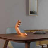 Copper finish geometric LED lamp by WOOM, radiating a warm glow on a wooden dining table.