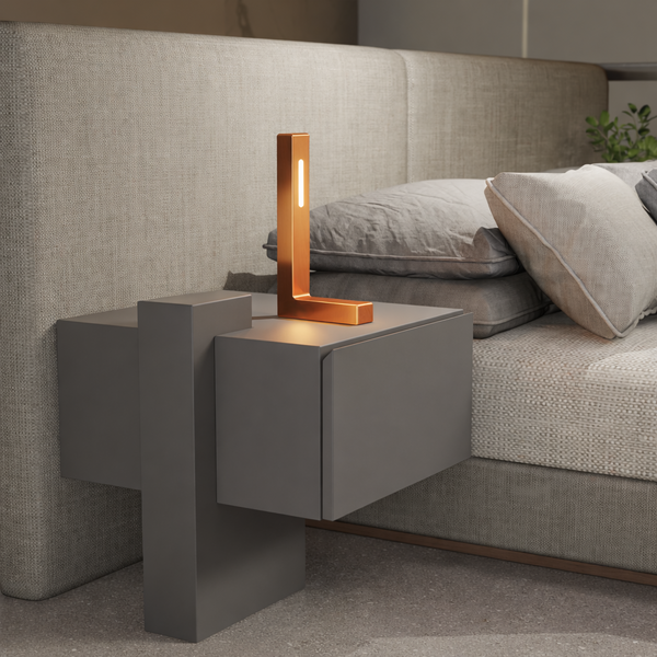 Copper finish L-shaped wooden lamp by WOOM on a dark gray nightstand next to a bed.