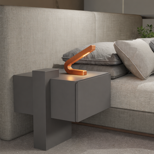 Handcrafted copper finish curved LED lamp by WOOM, casting a warm glow on a grey nightstand beside a bed.