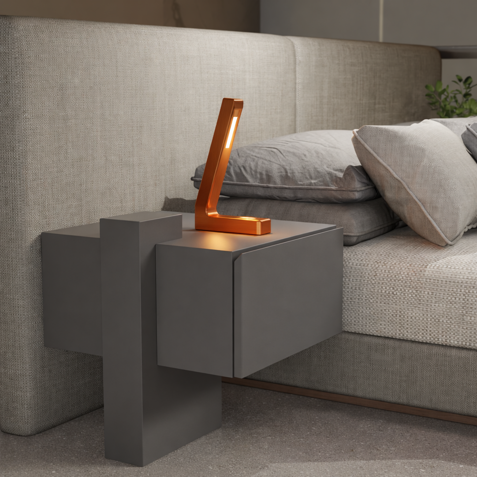 Copper finish L-shaped LED lamp by WOOM on a dark gray bedside table beside a bed.