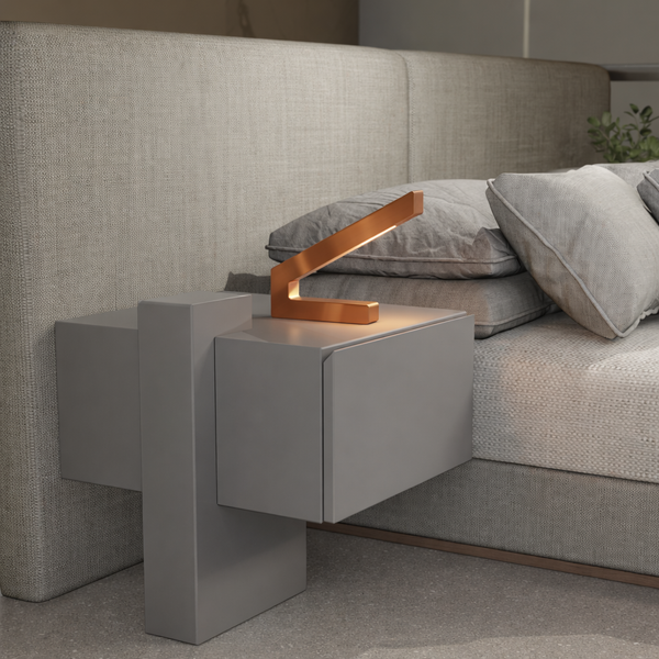 Copper finish modern LED lamp by WOOM with an angular design on a grey nightstand next to a fabric bed.