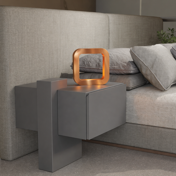 Square wooden lamp with a copper finish by WOOM, featuring an illuminated interior, on a grey nightstand beside a fabric bed.