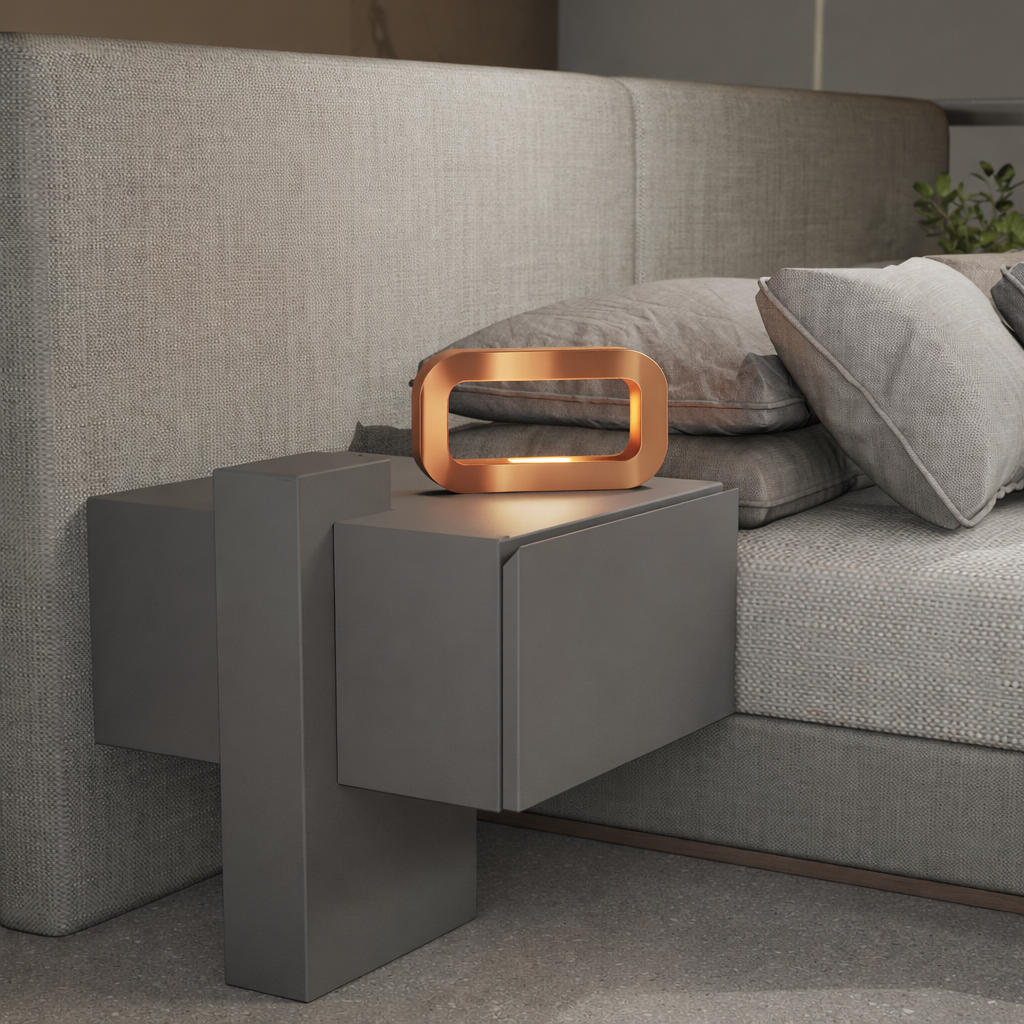 Copper finish rectangular table lamp by WOOM on a dark grey nightstand next to a light grey fabric bed.