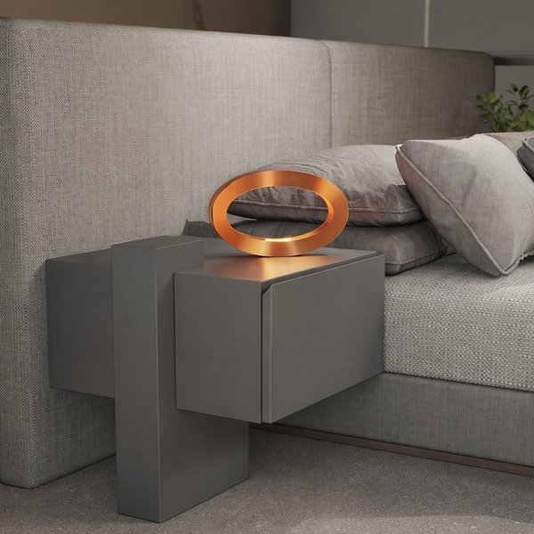Copper finish oval table lamp by WOOM, illuminating a dark grey bedside table next to a textured grey bed.