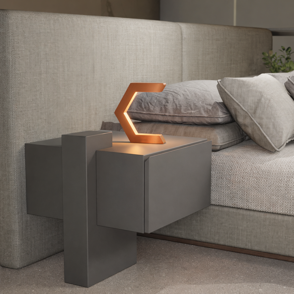 Copper C-shaped lamp by WOOM illuminating a modern gray nightstand beside a beige upholstered bed.