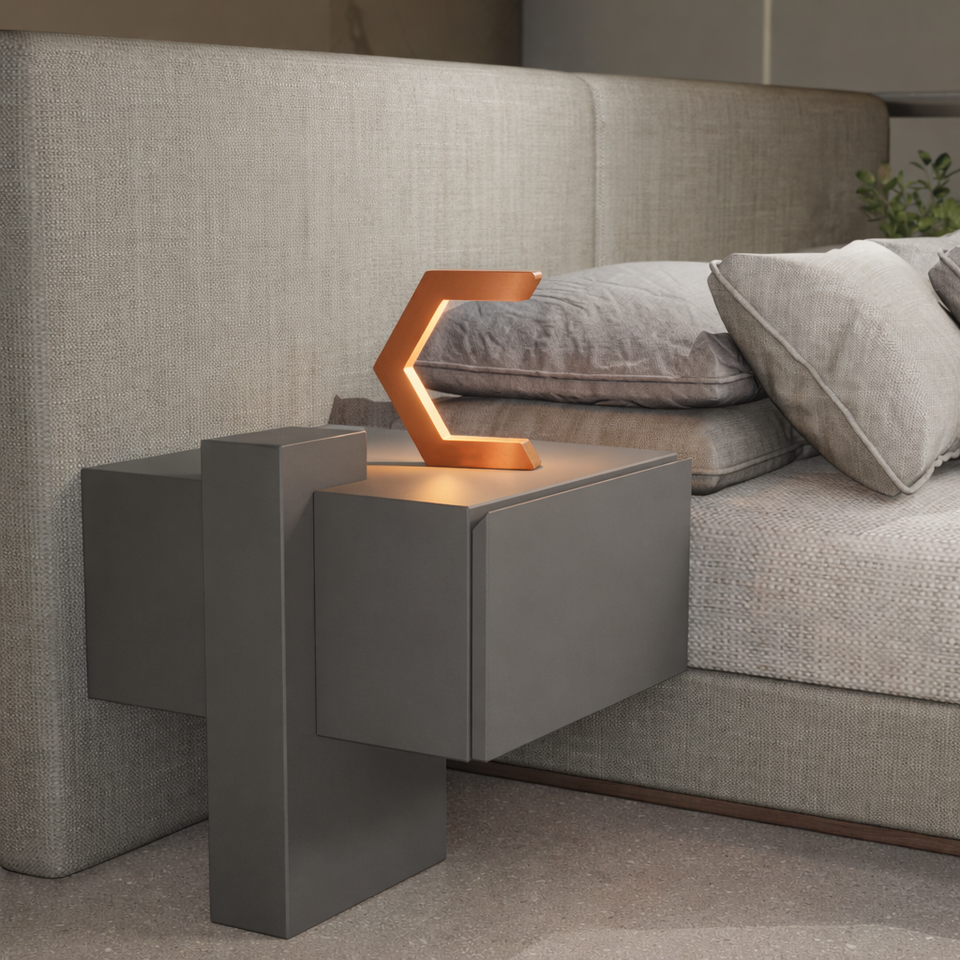 Copper C-shaped lamp by WOOM illuminating a modern gray nightstand beside a beige upholstered bed.