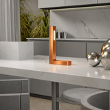 Copper finish oak L-shaped table lamp by WOOM, featuring a vertical glowing light strip, on a white marble counter.