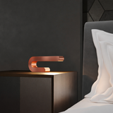 Copper finish U-shape curved lamp by WOOM, emitting a warm glow on a dark nightstand.