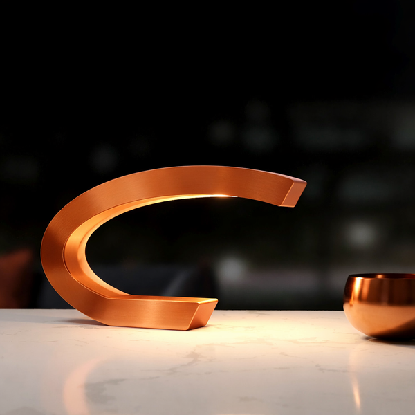 Copper finish C-shaped curved table lamp by WOOM, casting warm light on a marble surface next to a copper bowl.
