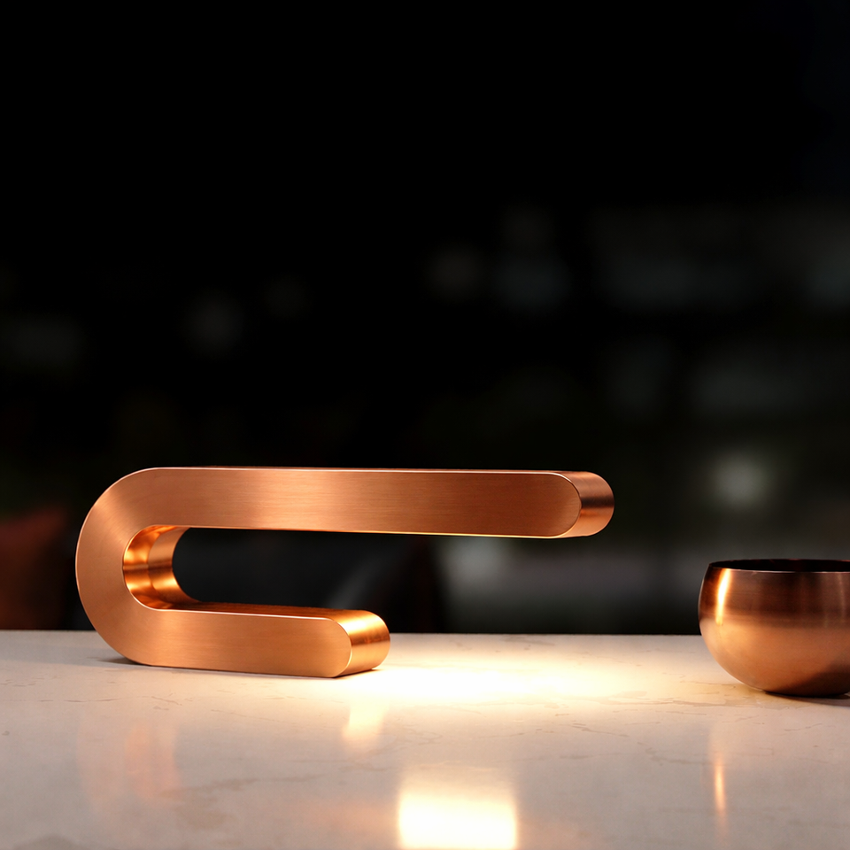 Copper finish U-shape curved lamp by WOOM, illuminating a light marble surface, next to a small copper bowl.