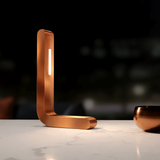 Copper finish L-shaped LED lamp by WOOM with a vertical light strip on a white marble countertop.