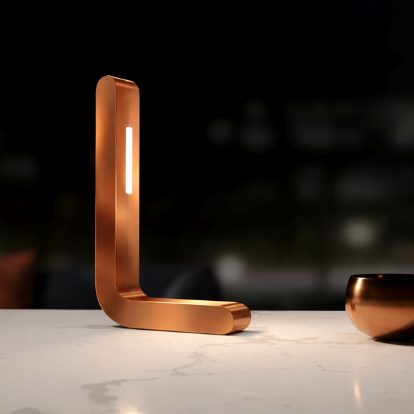 Copper finish L-shaped LED lamp by WOOM with a vertical light strip on a white marble countertop.