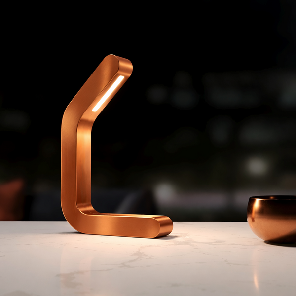 Handcrafted curved copper finish LED table lamp by WOOM, glowing on a white marble table next to a copper bowl.